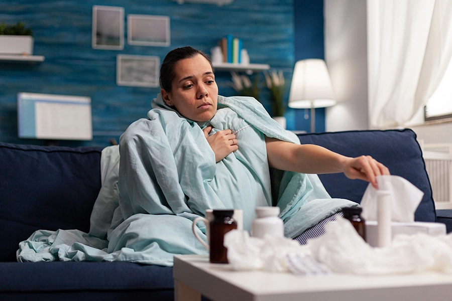 Sick woman sitting on couch, wrapped in a blanket, and reaching for a tissue